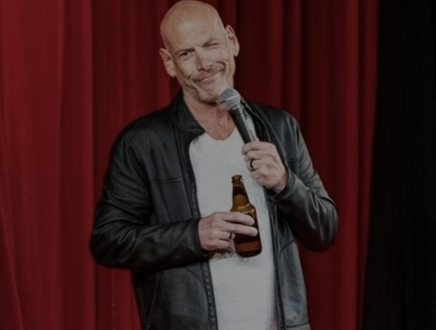 Bald man in a leather jacket holding a microphone and a beer bottle, standing in front of red curtains—looks like a comedian near me getting ready to perform.
